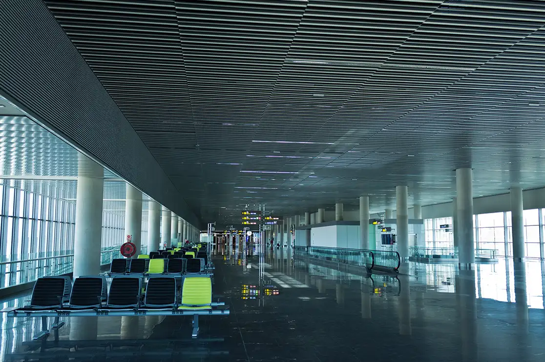 Airport passenger concourse with seating, a moving walkway, glazed facades, structural columns, and a suspended ceiling.