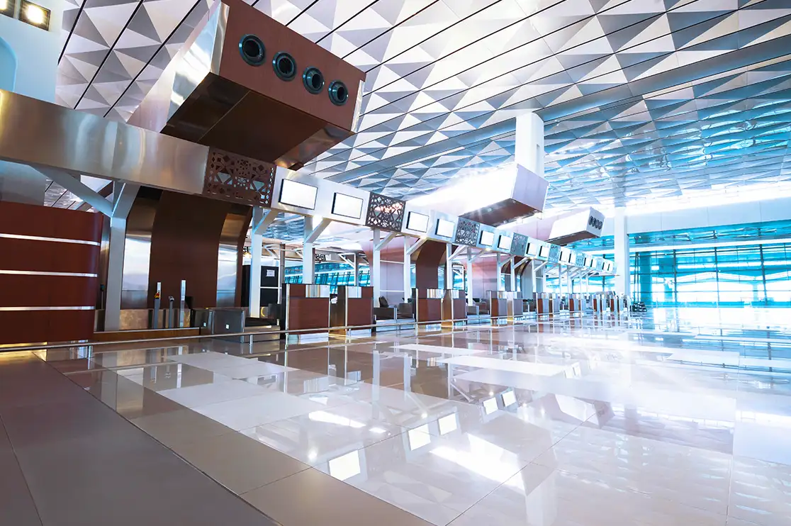 Modern airport terminal interior with a geometric ceiling, structural columns, glass facades, and overhead information displays