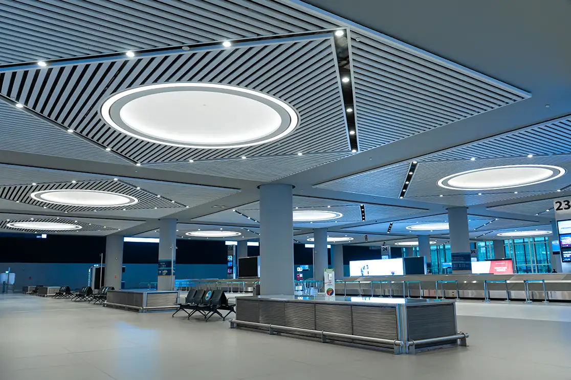 Airport terminal interior with a suspended linear ceiling, recessed lighting, structural columns, and passenger seating.