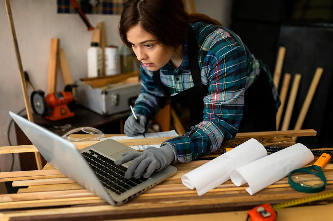 Millwork designer reviewing technical drawings on a laptop at a workshop table with woodworking materials, measuring tools, and rolled plans