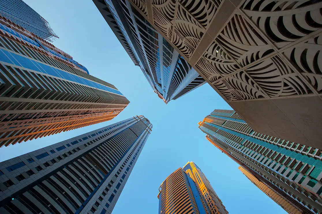 Worm’s-eye view of towering modern skyscrapers, highlighting their height, architectural facades, geometric details, and dramatic upward perspective against a clear blue sky.